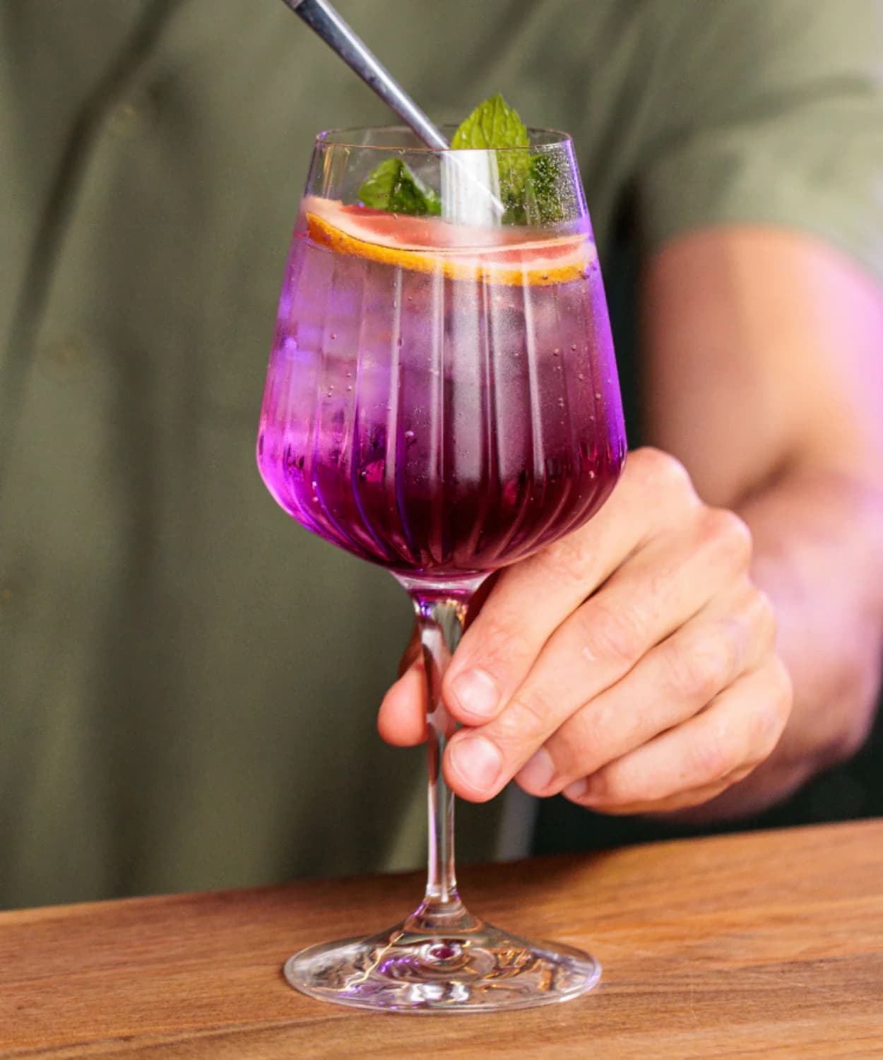 Cocktail viola in calice scanalato con pompelmo, menta e cannuccia metallica, tenuto da una mano.