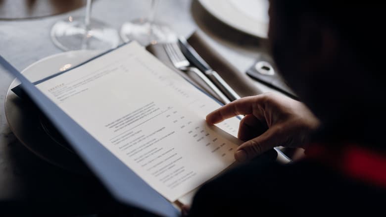 Mano che indica una voce sul menù di un ristorante elegante con tavola apparecchiata.